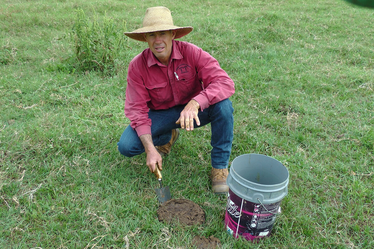 A man is crouching in a paddock with a small shovel and a bucket